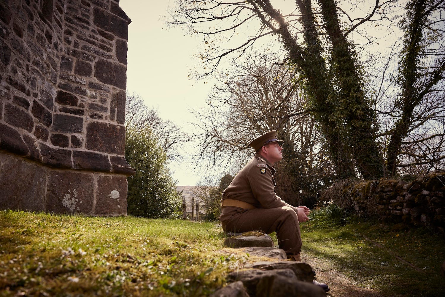 Tristan Farnon in uniform sits outside a farm building at the side of a track.