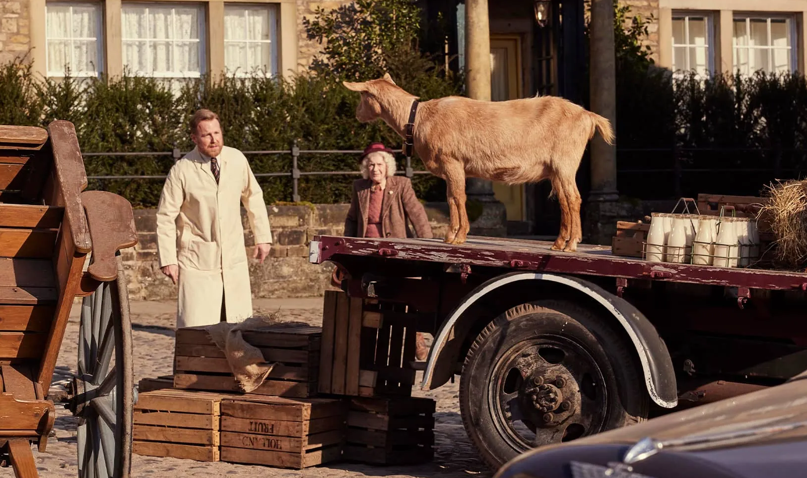 Hilda the goat stands on a cart carrying a crate of milk bottles outside Skeldale House. Siegfried Farnon attempts to catch her while her owner Mrs. Stokes watches.