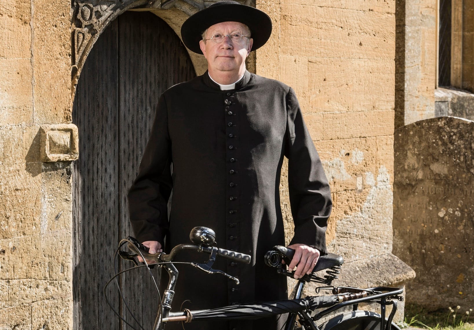 Father Brown (Mark Williams) outside his church with his bicycle.