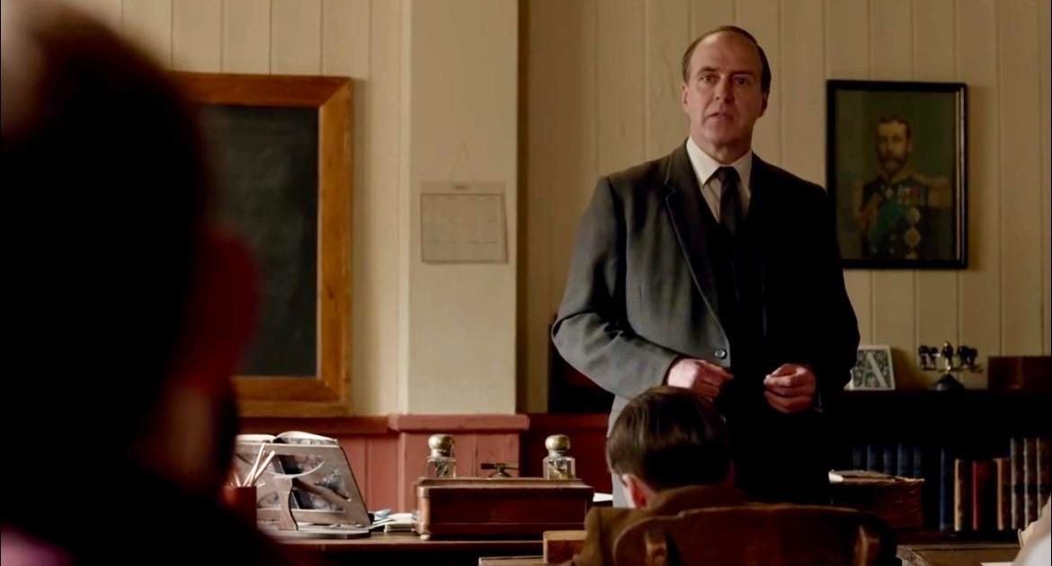 Mr. Molesley (Kevin Doyle) stands behind his desk
