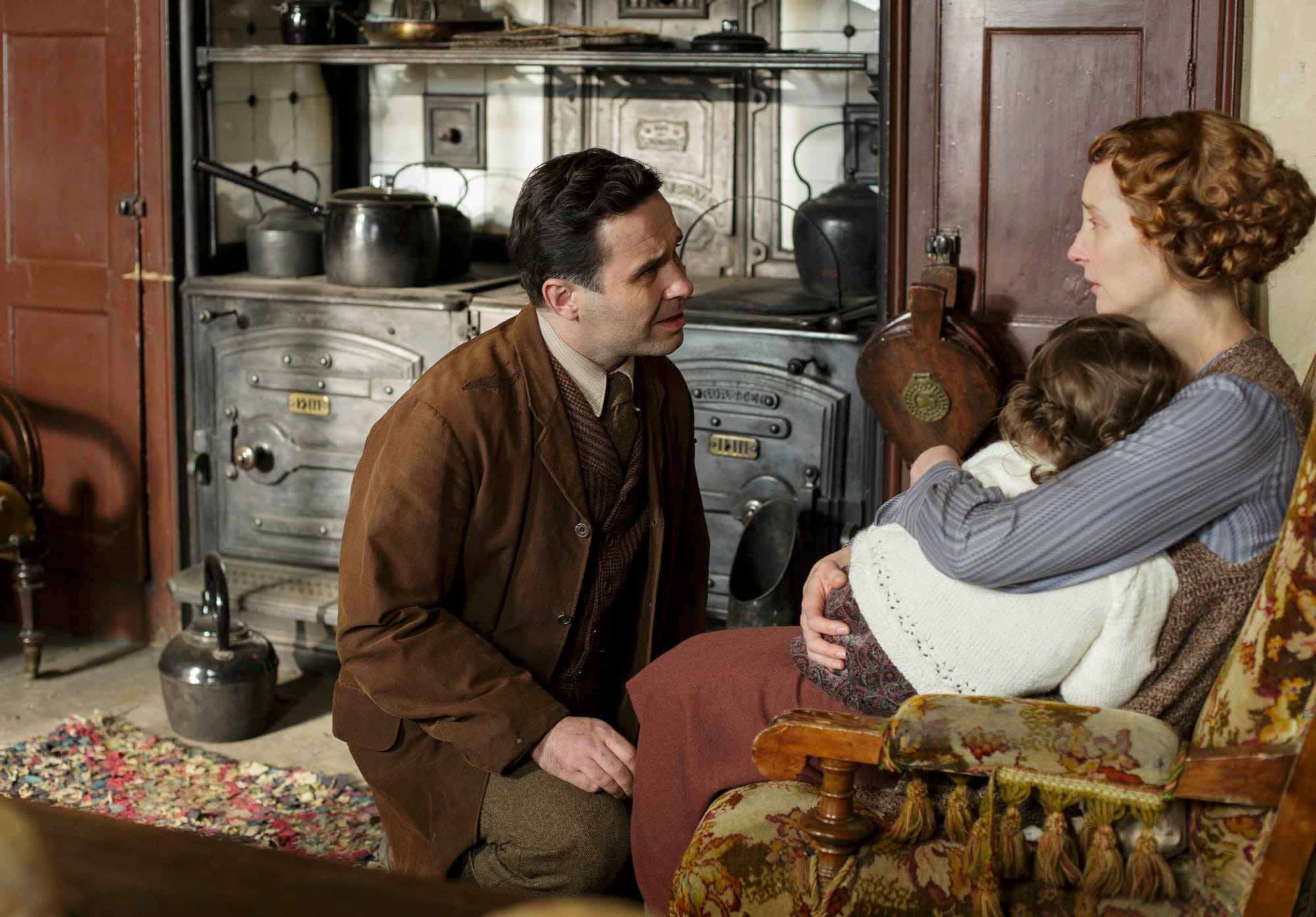 Tim Drewe (Andrew Scarborough) and his wife (Emma Lowndes), who cuddles Marigold (Eva Samms) seated in front of the kitchen range.