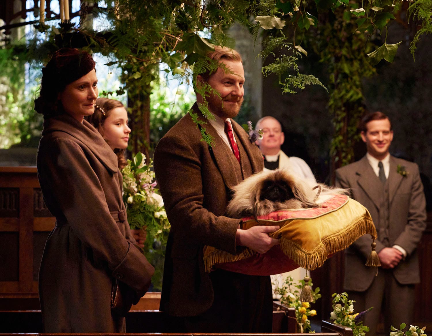 Mrs. Hall (Anna Madeley), Rachel Shenton (Imogen Clawsen), Siegfried Farnon (Samuel West), who holds the Pekinese Tricky Woo on a large pillow, and James Herriot (Nicholas Ralph) wait for the bride to enter the church.