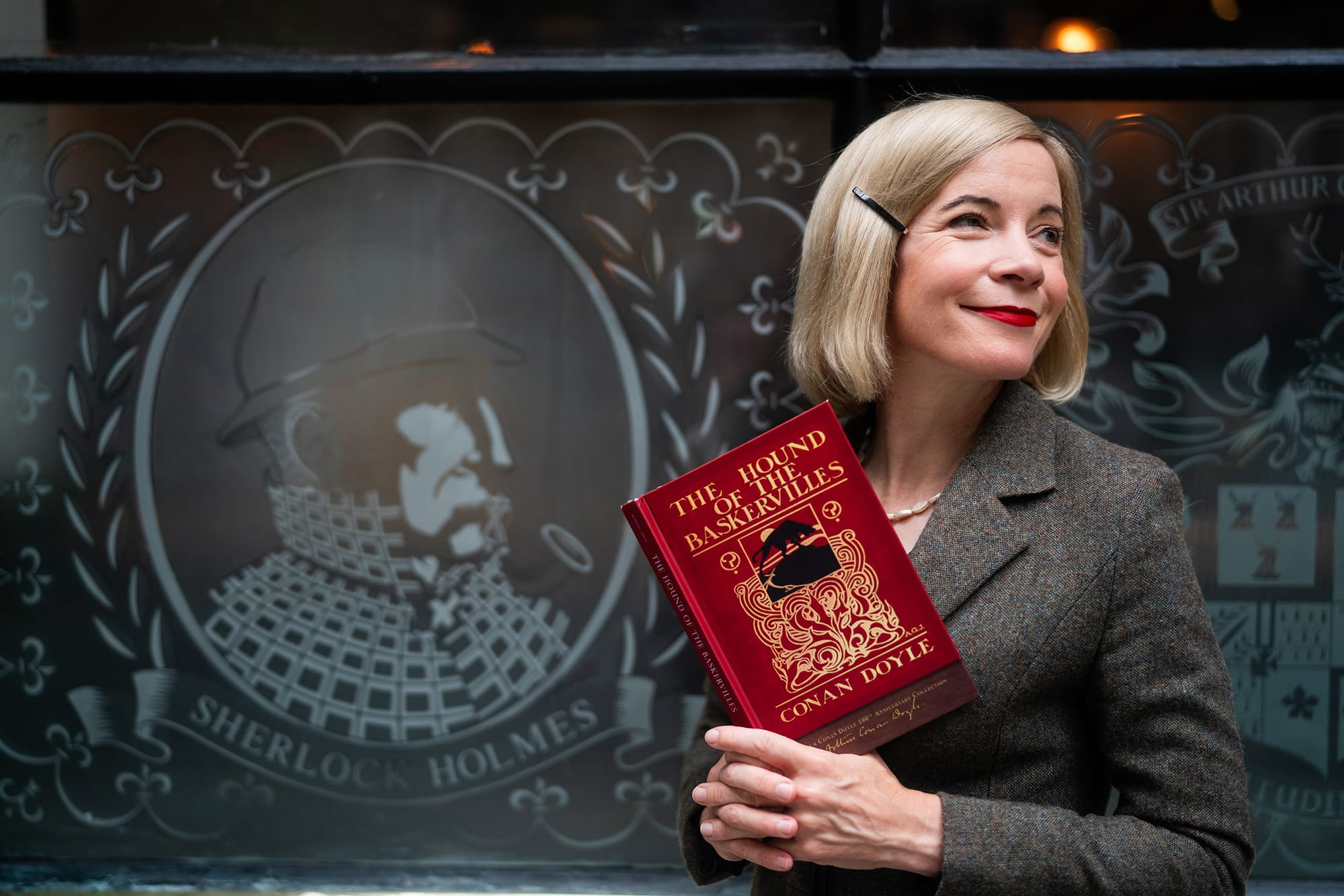 Lucy Worsley holding a copy of The Hound of the Baskervilles in front of The Sherlock Holmes Pub in London in 'Lucy Worsley's Holmes vs. Doyle'